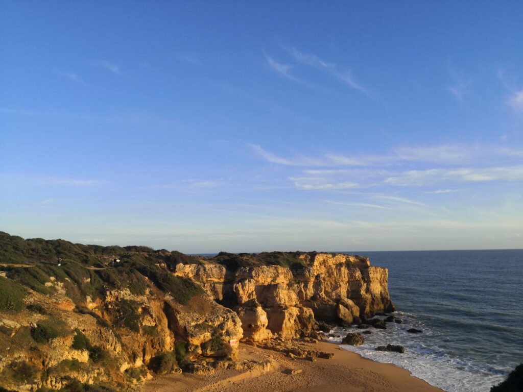Begrünte Klippen am Meer mit Sandstrand im Vordergrund. Blauer Himmel mit Federwolken.