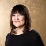 Frau mit schulterlangen, glatten, dunklen Haaren und schwarzem Oberteil lächelt