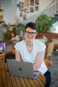 Olivia trägt ein weißes Top, Brille, lächelt und sitzt vor einem Laptop
